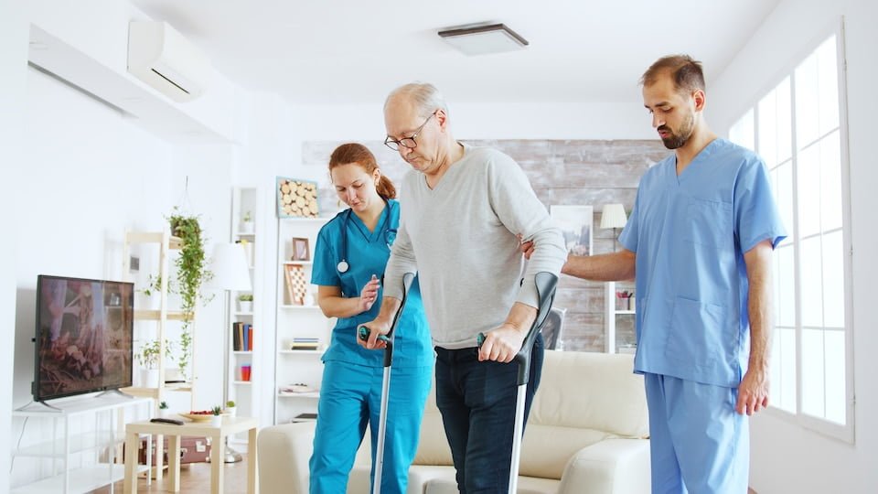 Female doctor and her assistant helping old woman with crutches to stand up from the couch and take a walk